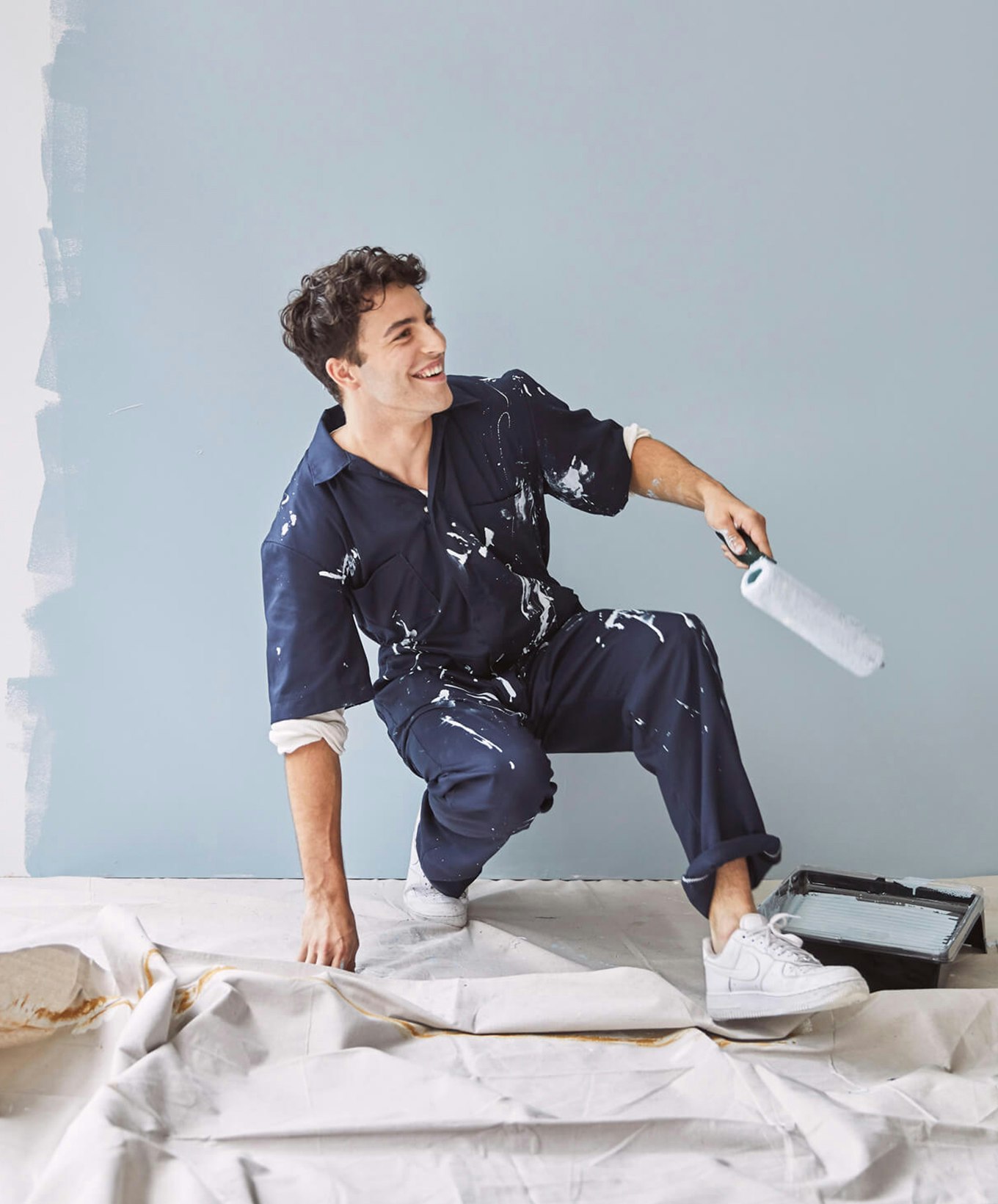 Man in a dark blue jumpsuit holding a roller brush crouching in front of a painted wall