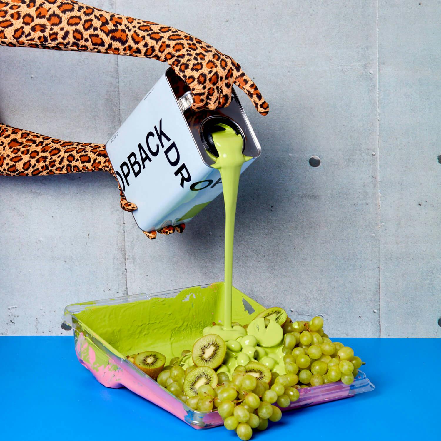 A can of PRETTY UGLY paint being poured onto green grapes and kiwi by hands wearing leopard-print gloves