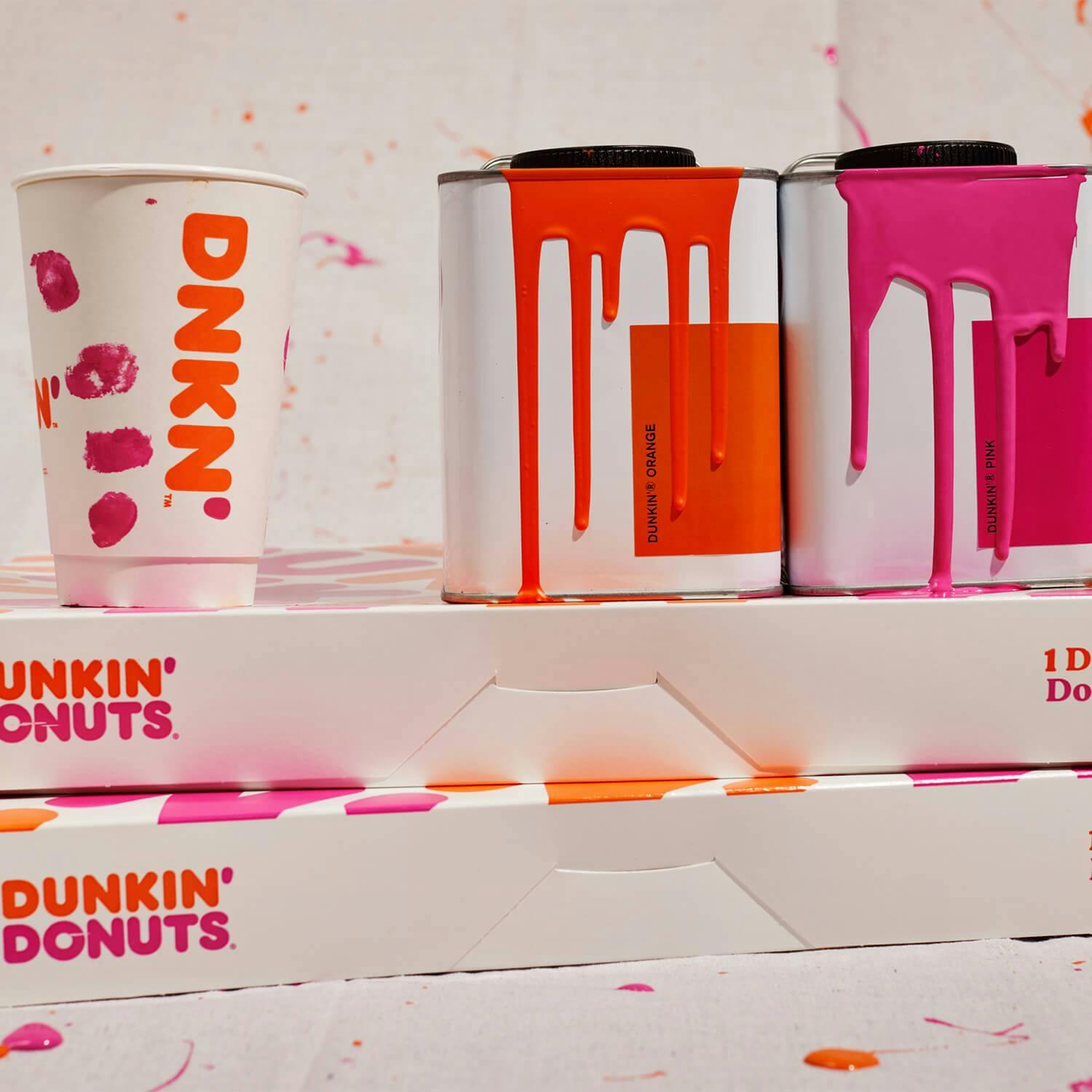 Dunkin'® Orange and Dunkin'® Pink paint cans next to a coffee cup on top of boxes of donuts