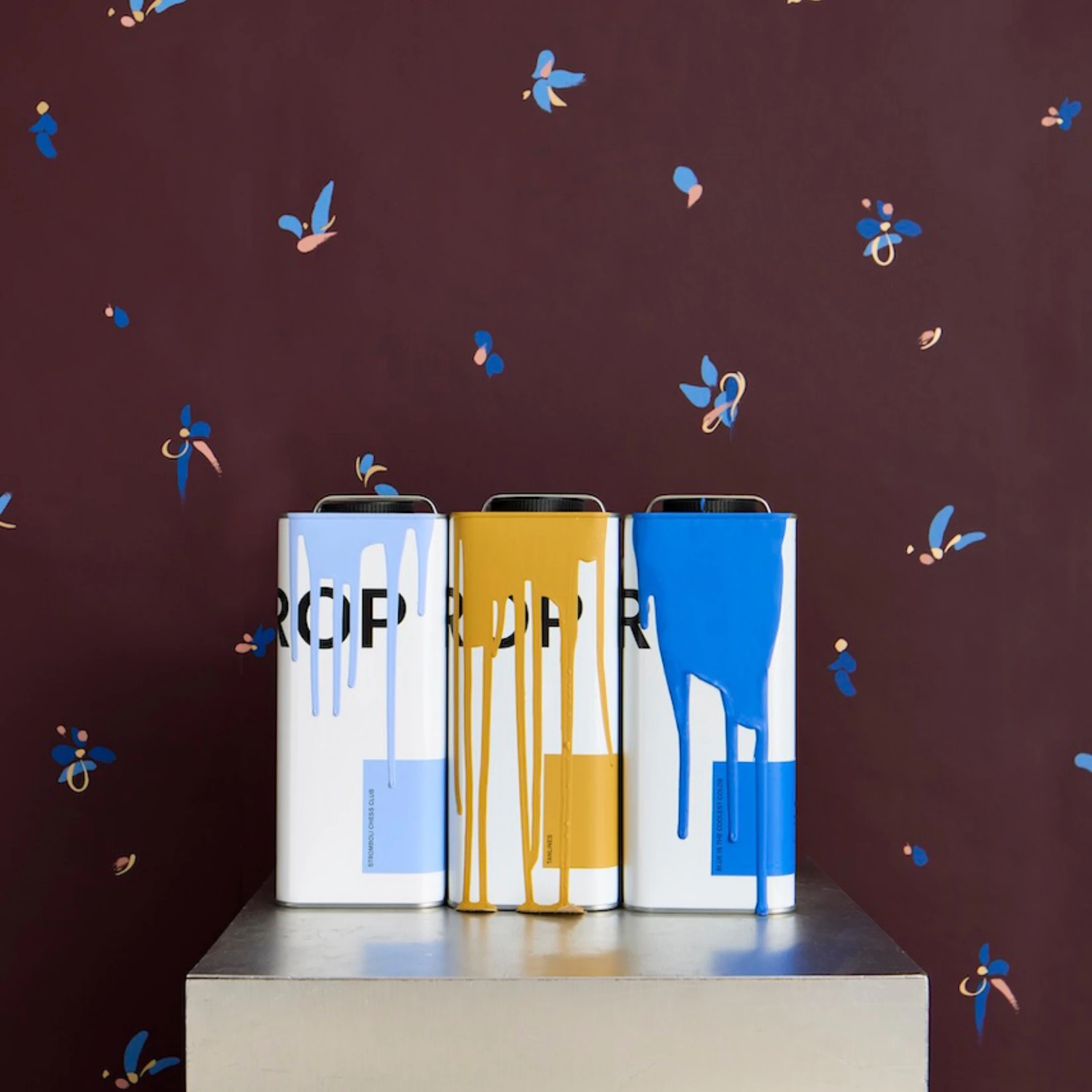 Three paint cans sit on top of a chrome pillar in front of Backdrop's LA BELLE wallpaper in Warm Purple