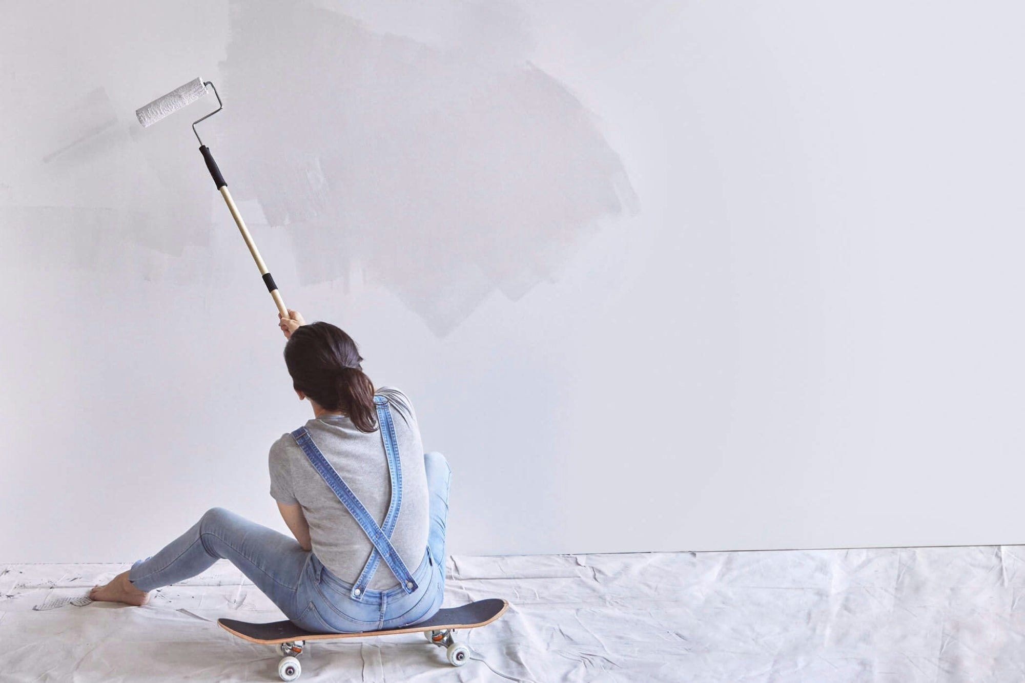 Person sitting on a skateboard and painting a wall with a paint roller with extension handle