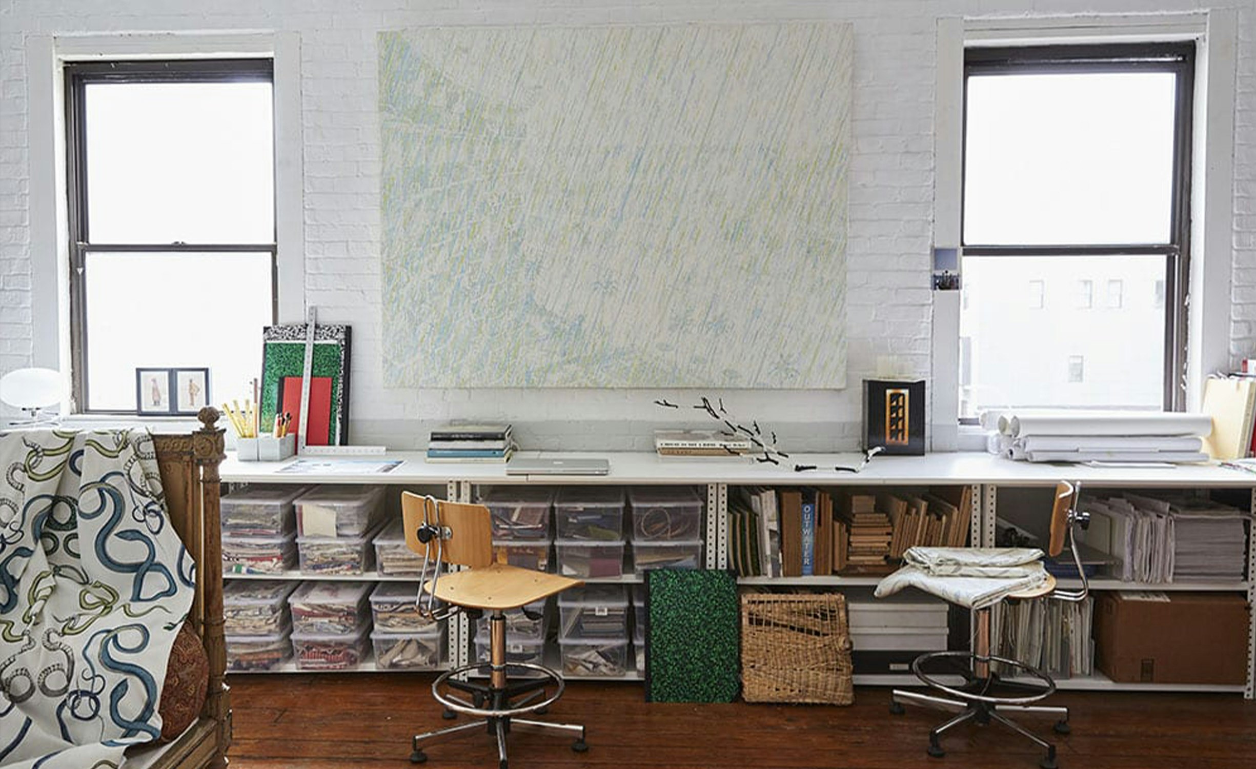 A room filled with natural light shows a long desk in front of a large piece of art between two windows