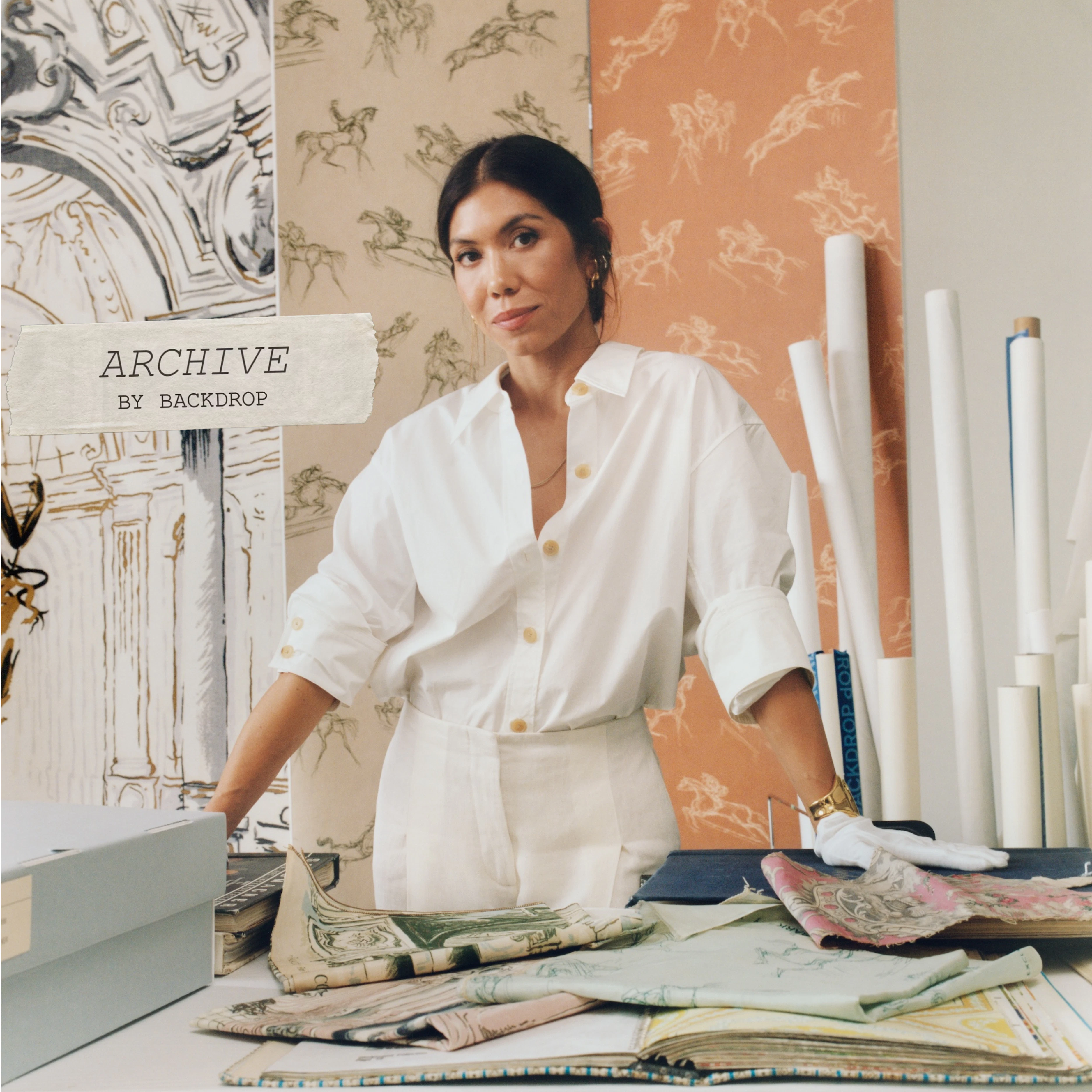 Backdrop's founder, Natalie Ebel, wearing white archivist gloves and looking through fabric and wallpaper samples from the Schumacher Archive that inspired ARCHIVE by BACKDROP, a new wallpaper collection
